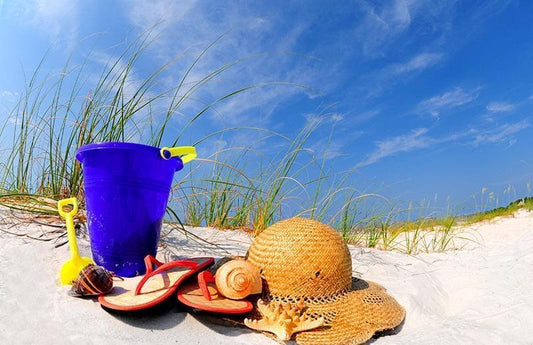 Low angle view of blue bucket, sunhat, flip flops, and sea shells on white sand beach with cloudless sky background as a visual representation of Beach Day Fragrance Oil available at Village Craft and Candle 