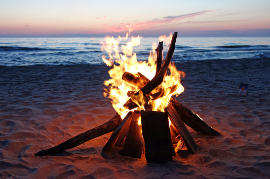  Feu de camp brûlant sur une plage au coucher du soleil comme représentation visuelle de l'huile parfumée de feu de camp disponible chez Village Craft and Candle