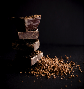 Four pieces of dark chocolate stacked on top of each other against a black background with a pile of chocolate shavings spread before it || Quatre morceaux de chocolat noir empilés les uns sur les autres sur un fond noir avec un tas de copeaux de chocolat répartis devant lui