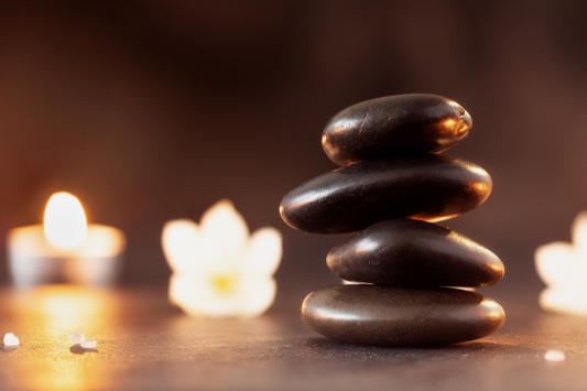 Stacked brown stones in foreground with flowers and candles as a visual representation of Spa Fragrance Oil available at Village Craft and Candle 