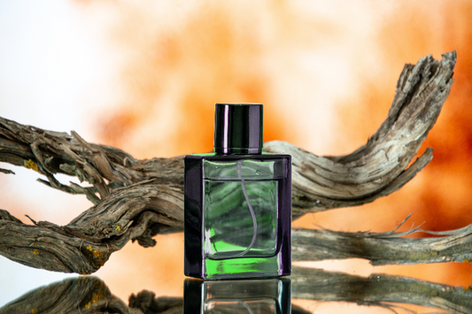 Green glass cologne bottle sitting on mirrored surface in front of driftwood on an orange & gold background as a visual representation of Kristoff 1972 Fragrance Oil available at Village Craft and Candle 