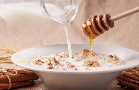 Bowl of oatmeal with milk and honey pouring in, next to cinnamon sticks and wheat as a visual representation of Oatmeal and Warm Honey Fragrance Oil available at Village Craft and Candle 