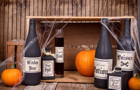 Black bottles of poison covered in cobwebs and surrounded by pumpkins and wooden crates as a visual representation of Witches Stew (Black Licorice) Fragrance Oil available at Village Craft and Candle 